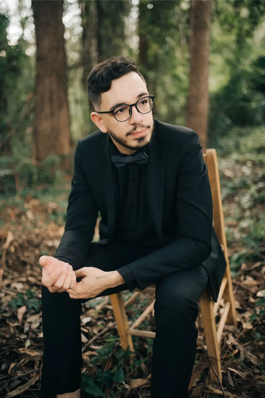 Noivo de &oacute;culos vestido de terno preto completo com gravata borboleta, sentado em meio &agrave; natureza.