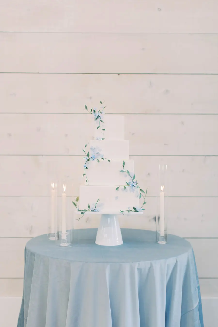 Bolo de casamento branco de quatro andares decorado com flores delicadas azuis, sobre mesa com toalha azul.