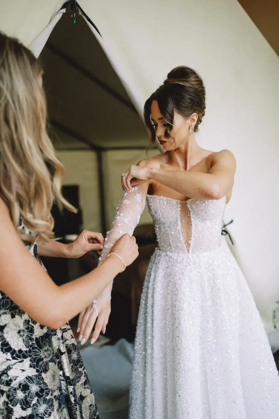 Noiva ajustando vestido de mangas longas com ajuda de madrinha, usando coque alto elegante.