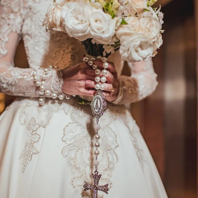 Noiva segurando buquê de rosas brancas com terço de pérolas e medalha prateada.