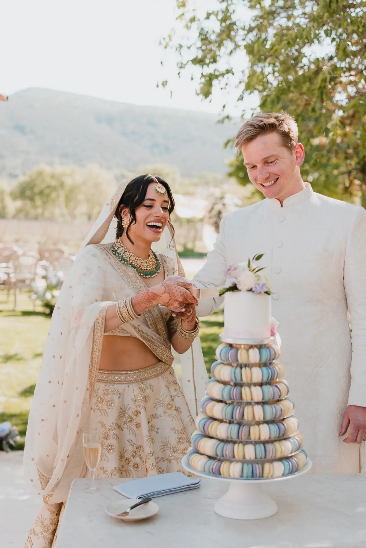 Casal de noivos sorridente cortando bolo com torre de macarons coloridos ao ar livre.