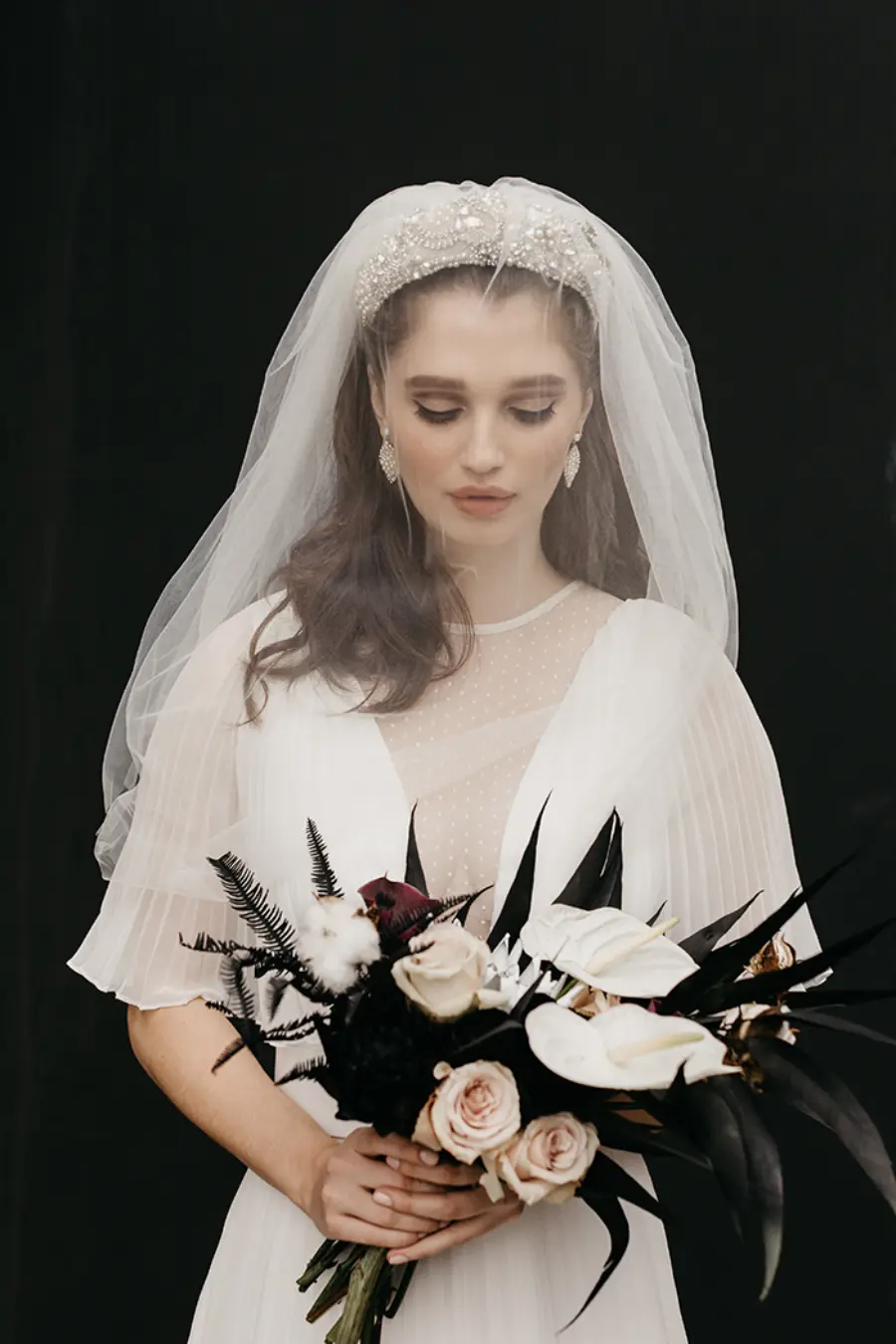 Noiva com v&eacute;u e tiara de p&eacute;rolas segurando buqu&ecirc; elegante com rosas claras e flores brancas contrastando com folhas escuras.