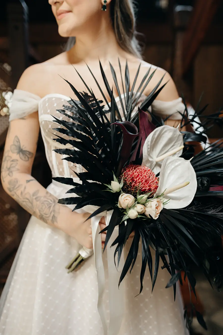 Noiva sorridente segurando um buqu&ecirc; moderno com folhas pretas, flores ex&oacute;ticas brancas e detalhes vermelhos.