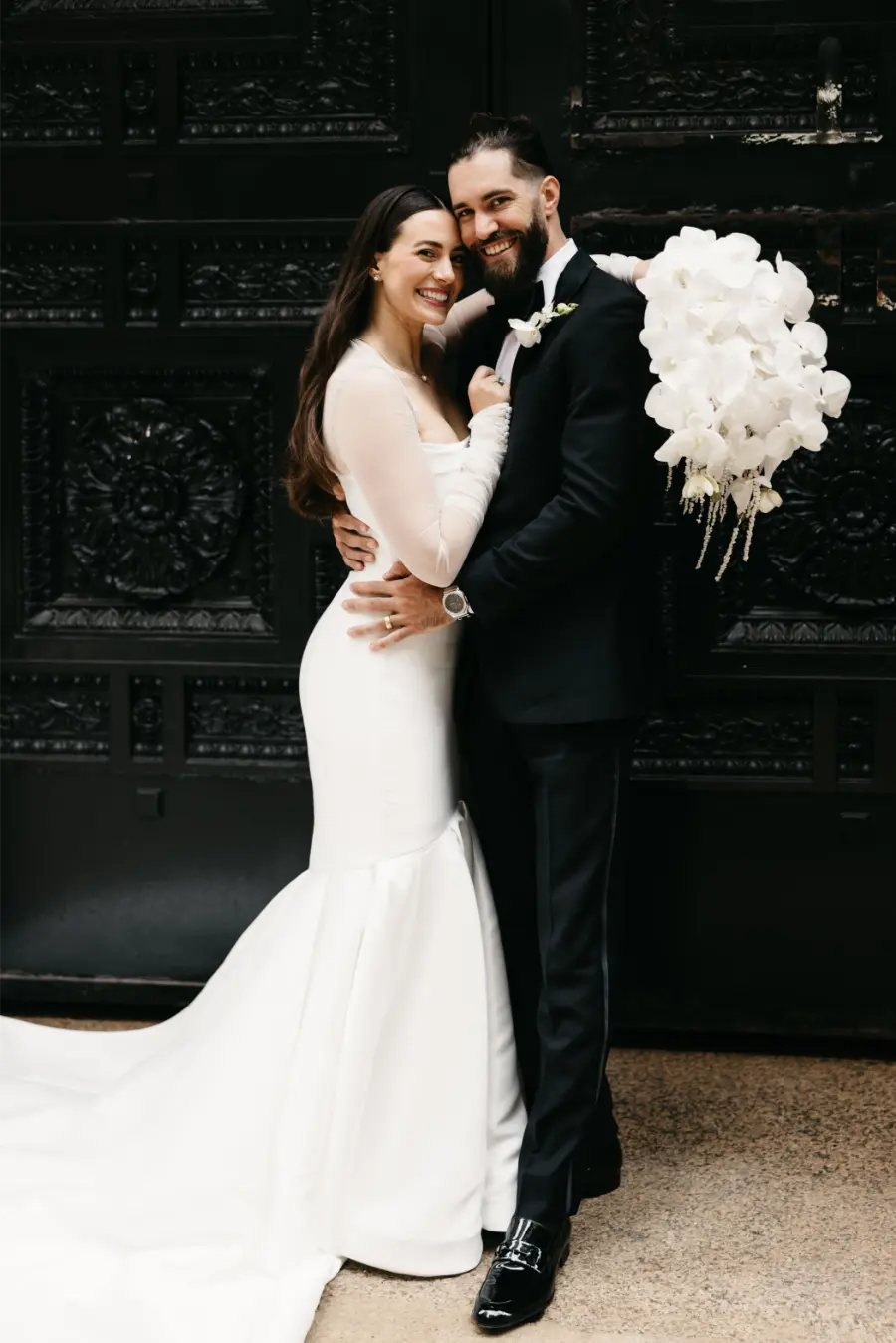 Casal de noivos abra&ccedil;ados sorrindo em frente a port&atilde;o ornamentado preto, noiva segura buqu&ecirc; de orqu&iacute;deas brancas