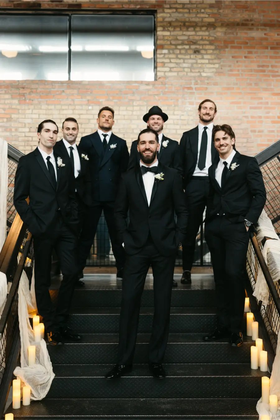 Padrinhos de casamento posando em escada, todos de terno preto com boutoni&egrave;res, em cen&aacute;rio com velas decorativas.