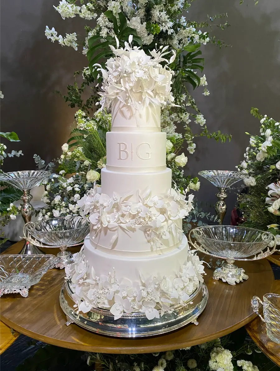 Bolo de casamento branco de cinco andares com detalhes em flores brancas delicadas ao redor, apoiado em base espelhada e com iniciais “B G” no centro.