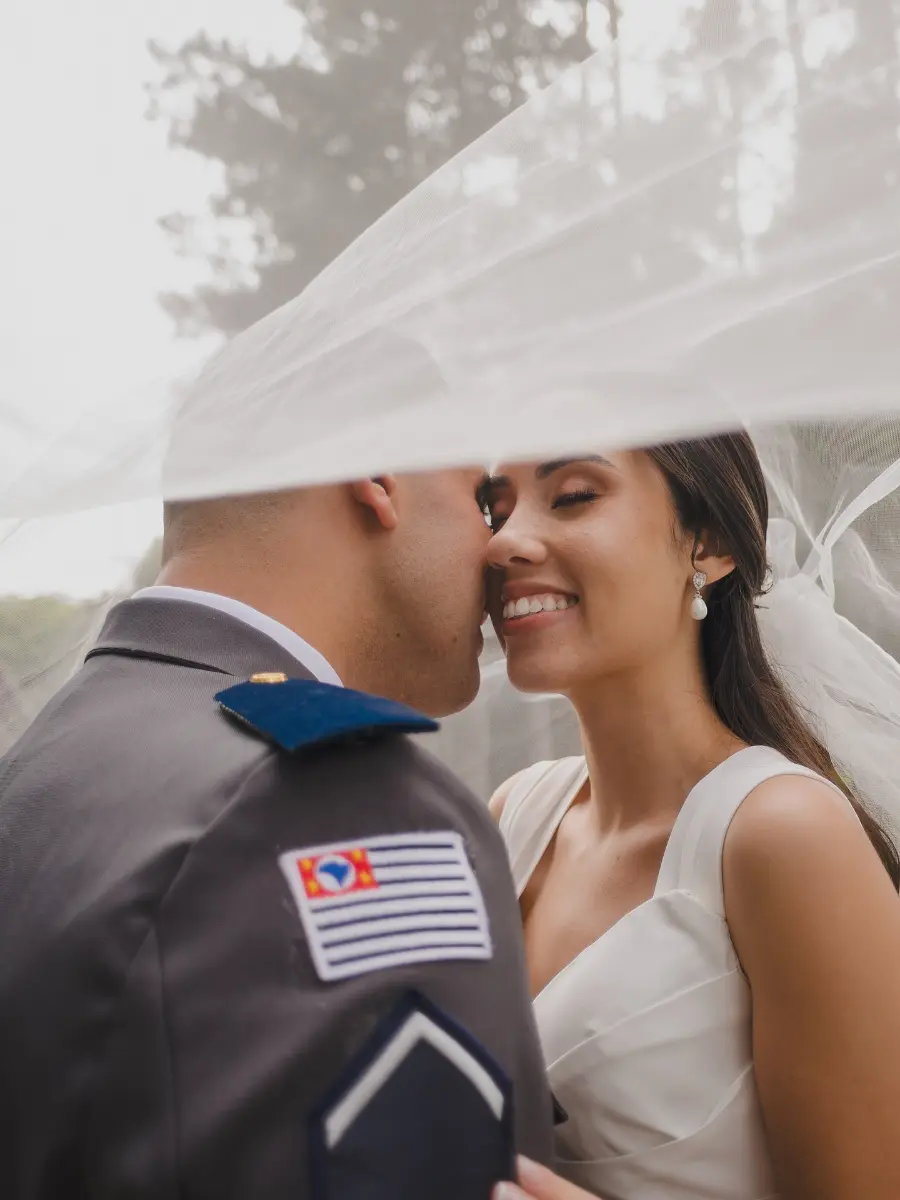 Casal apaixonado em casamento militar, noivo com uniforme de gala e noiva de vestido branco sob v&eacute;u leve.