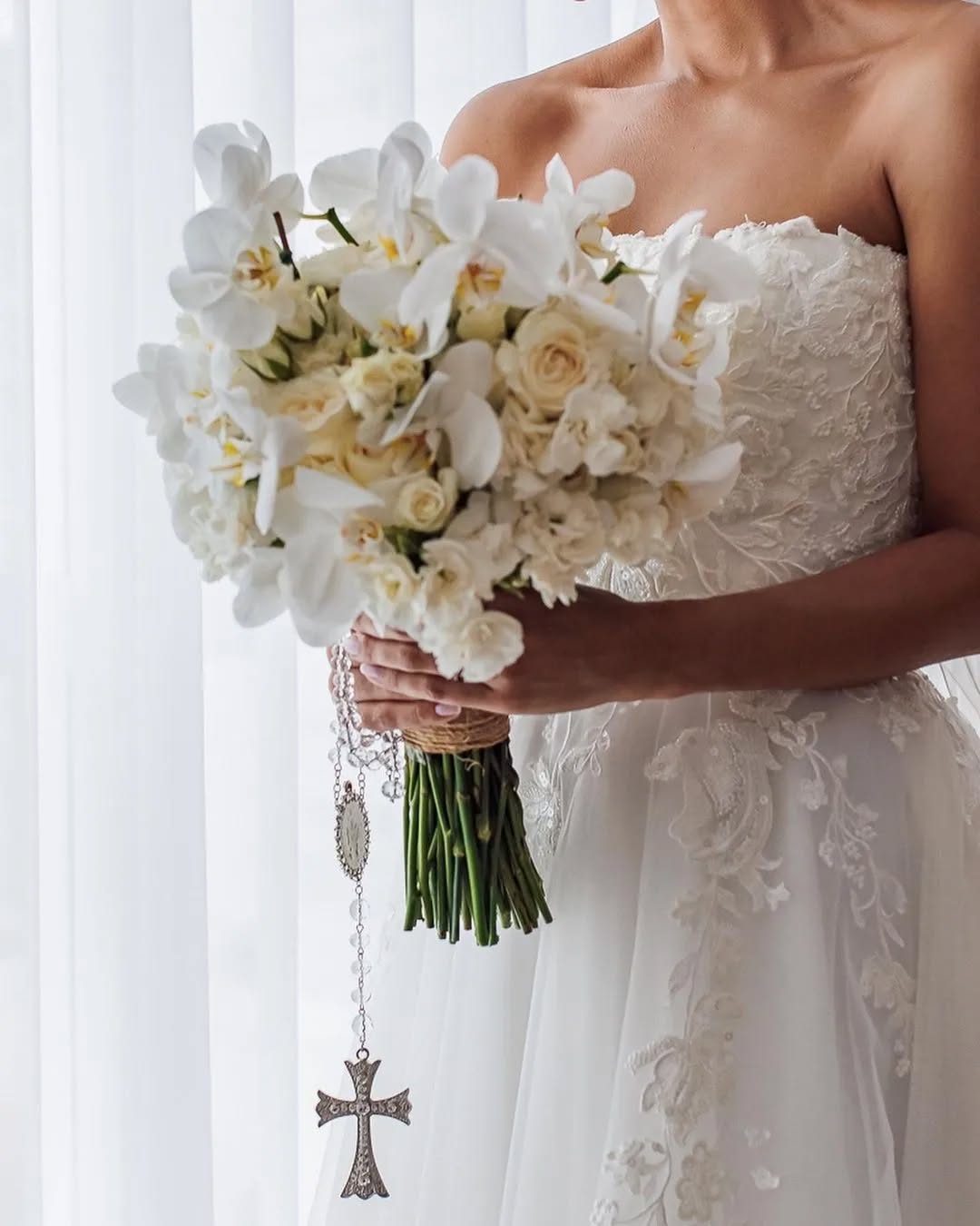 Noiva segurando buquê branco com terço prateado pendurado.