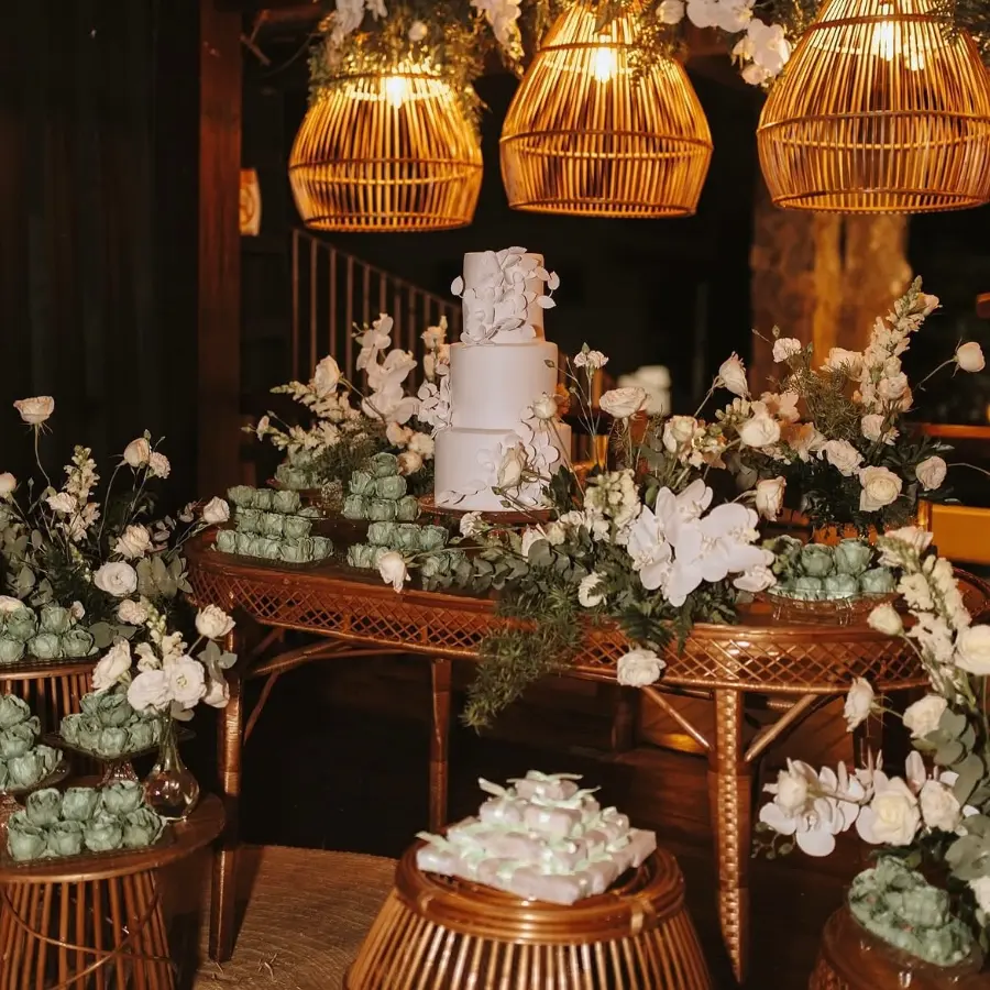 Mesa de casamento decorada com bolo fake branco de três andares, flores brancas e doces verdes em ambiente rústico
