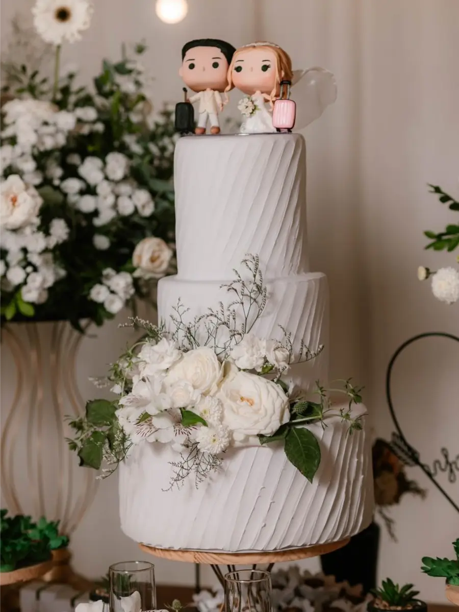 Bolo branco de três andares com textura diagonal, decorado com flores brancas naturais e topo com bonecos de noivos estilizados.