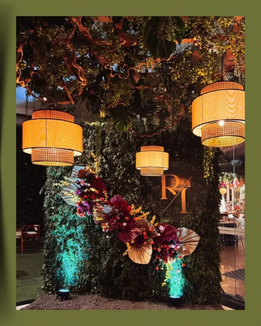 Painel de casamento com folhagens verdes, flores coloridas e monograma dourado "R/I", iluminado por luminárias suspensas.