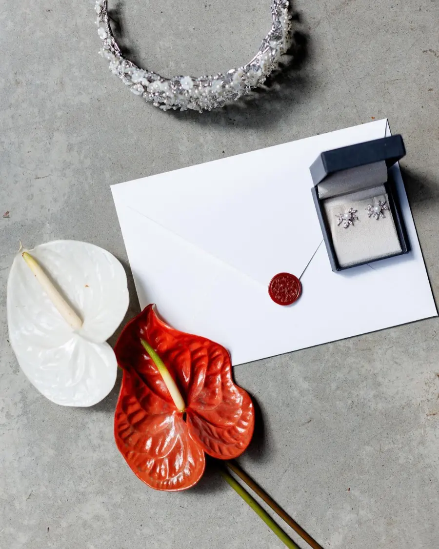 Flat lay de papelaria de casamento com envelope lacrado, brincos de diamante, flores antúrio vermelhas e vela sobre fundo cinza