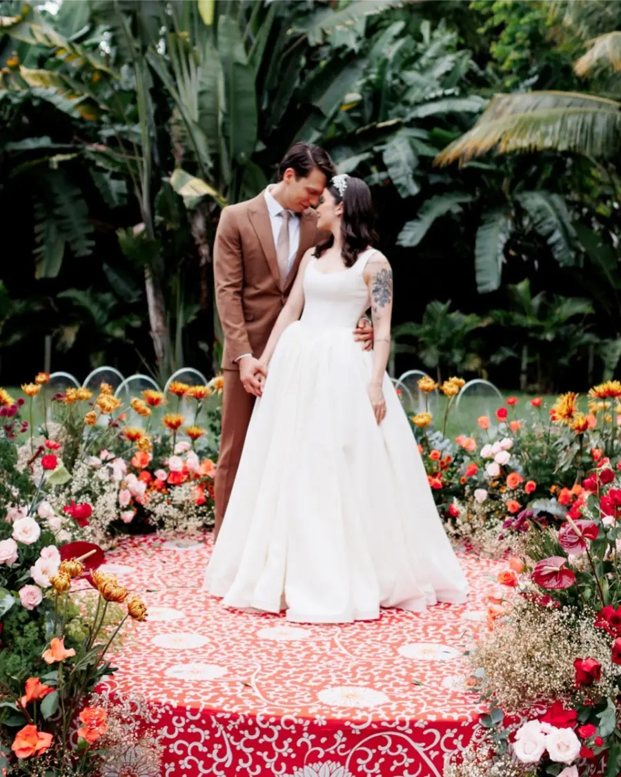 Casal de noivos se beijando em cerimônia ao ar livre com tapete vermelho florido e jardim tropical ao fundo