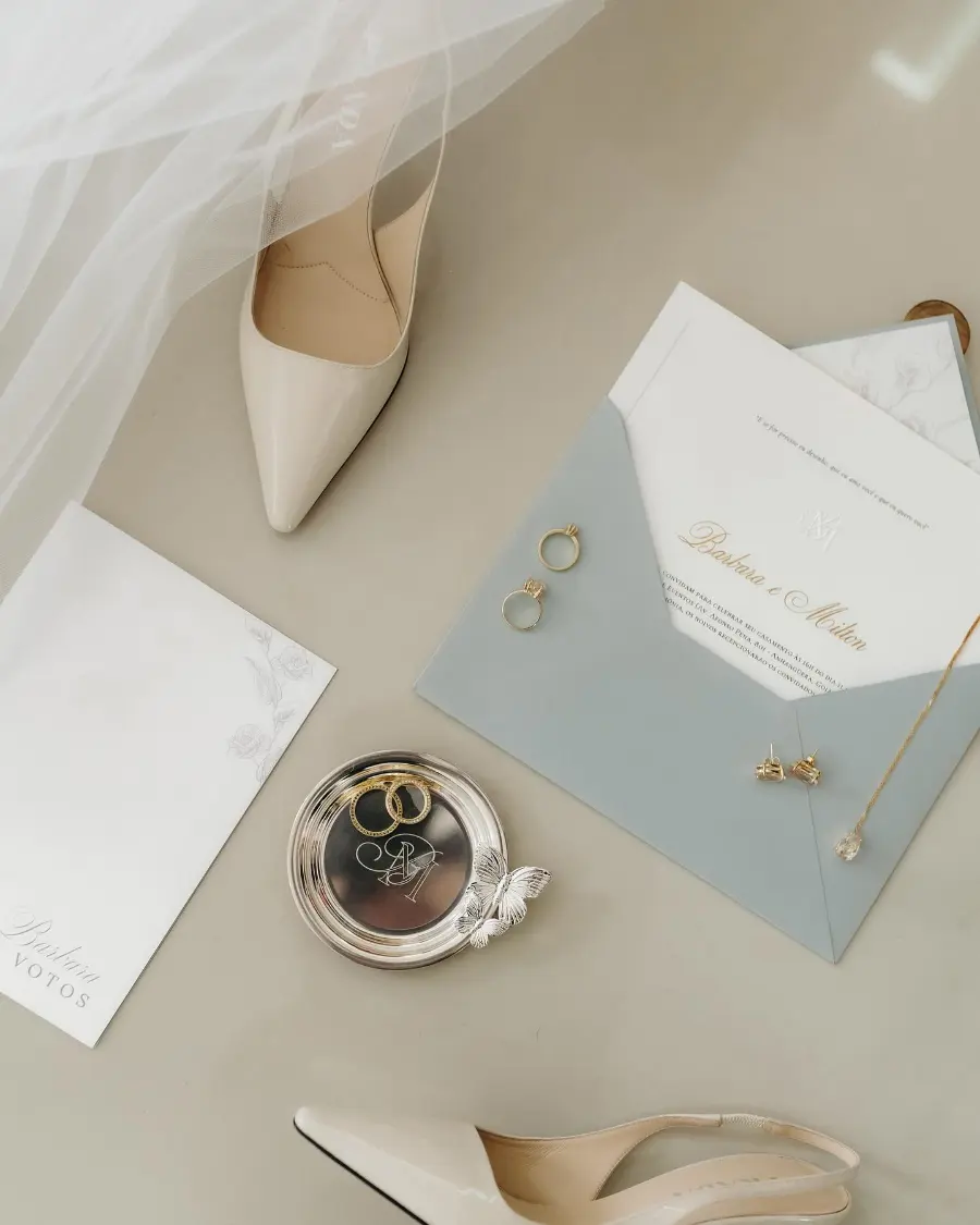 Flat lay de acessórios de noiva com sapatos bicos brancos, alianças douradas, convite e véu sobre fundo neutro