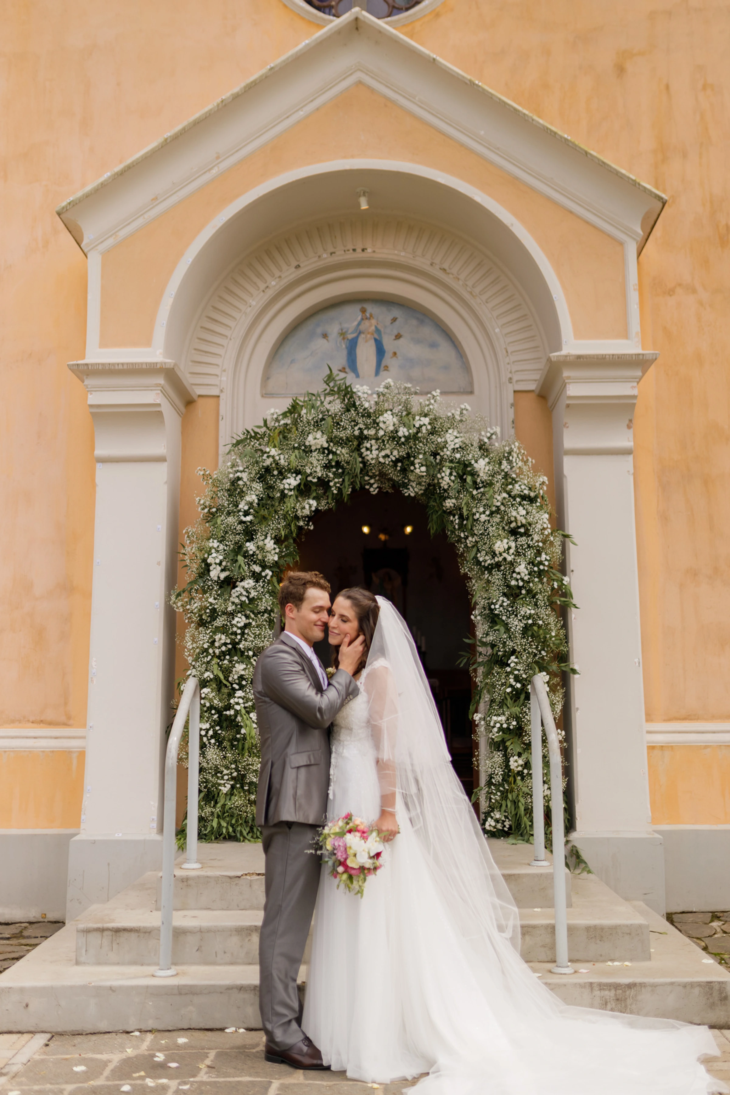 Noivos posando na entrada de capela com fachada amarela e arco de flores brancas
