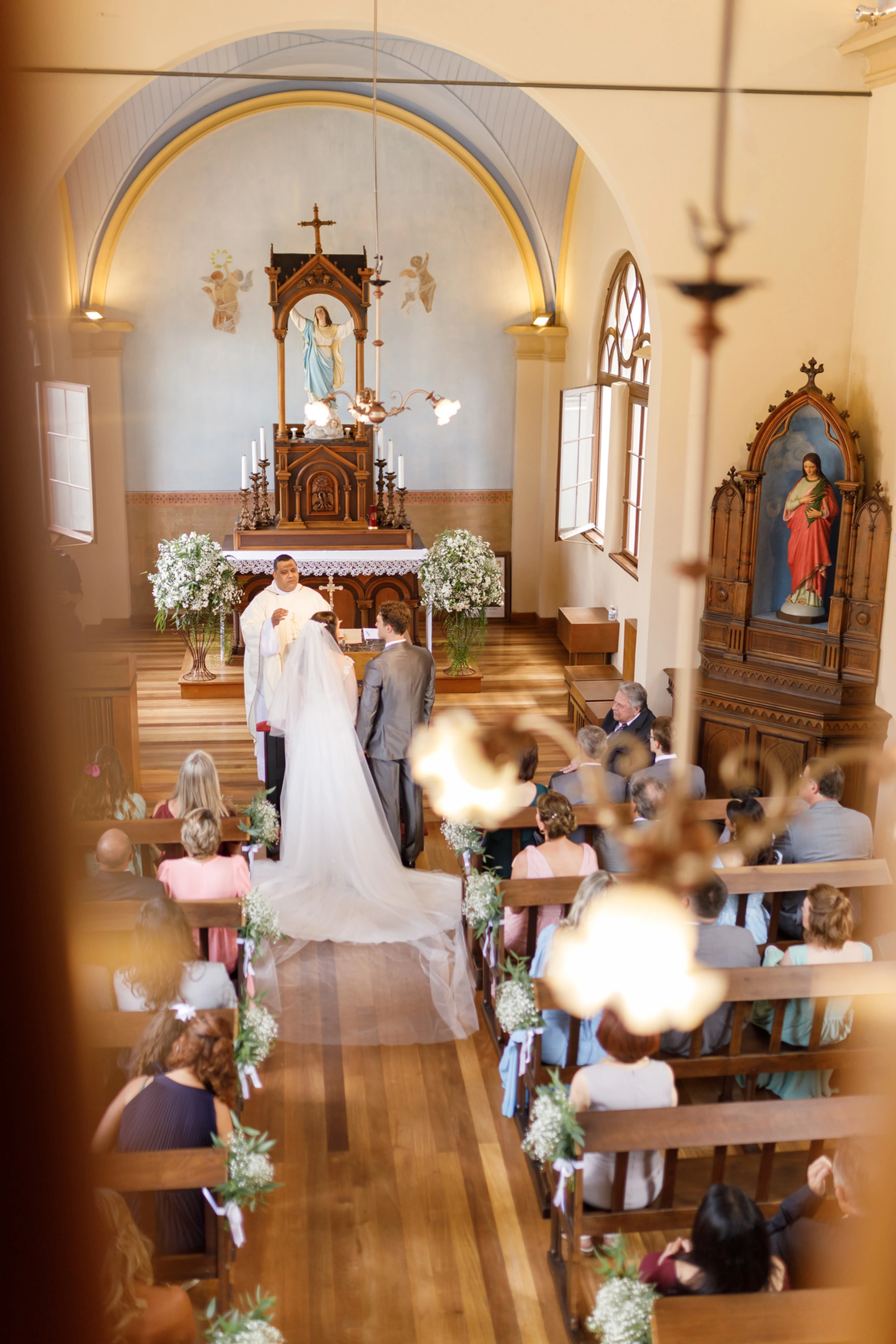 Noivos no altar de capela tradicional com bancos de madeira e flores brancas