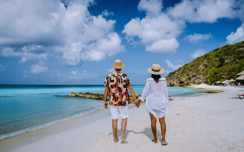 casal de costas caminhando de m&atilde;os dadas em uma praia tropical de areia branca e &aacute;guas cristalinas.