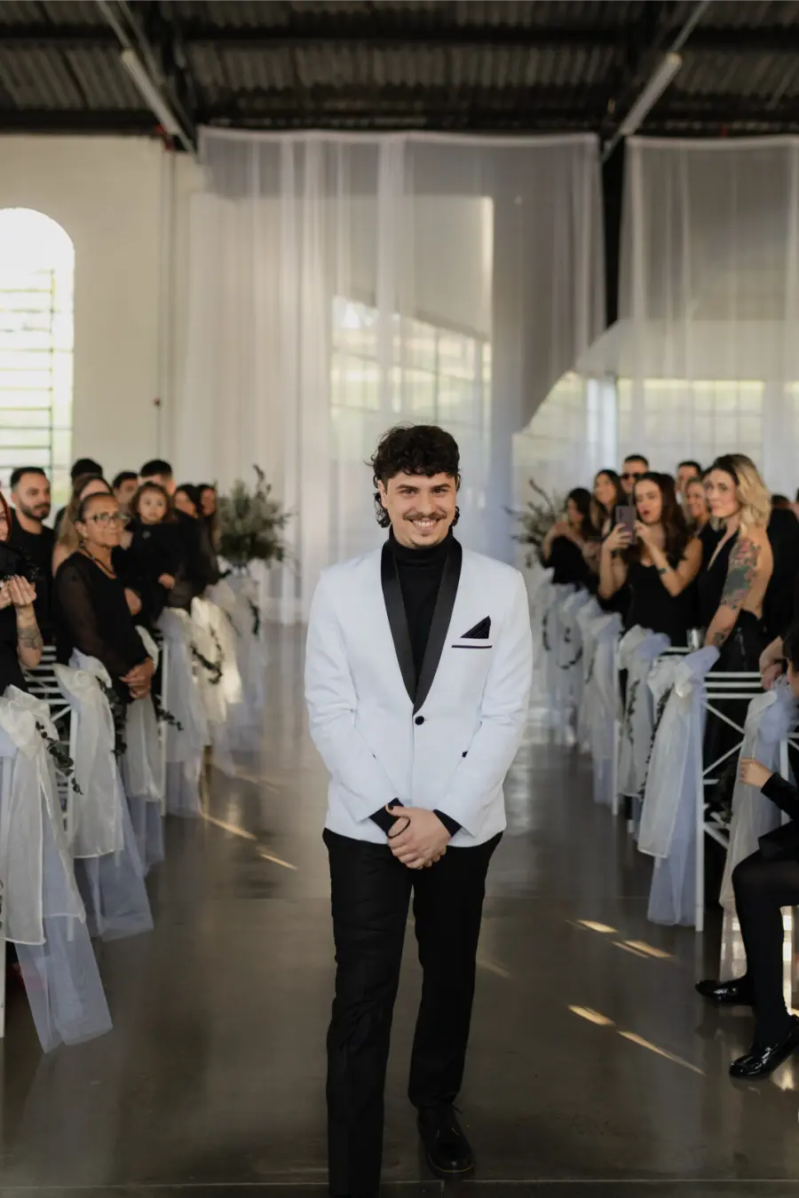 Noivo sorridente desfilando no corredor da cerim&ocirc;nia, usando palet&oacute; branco com gola preta e camisa escura.
