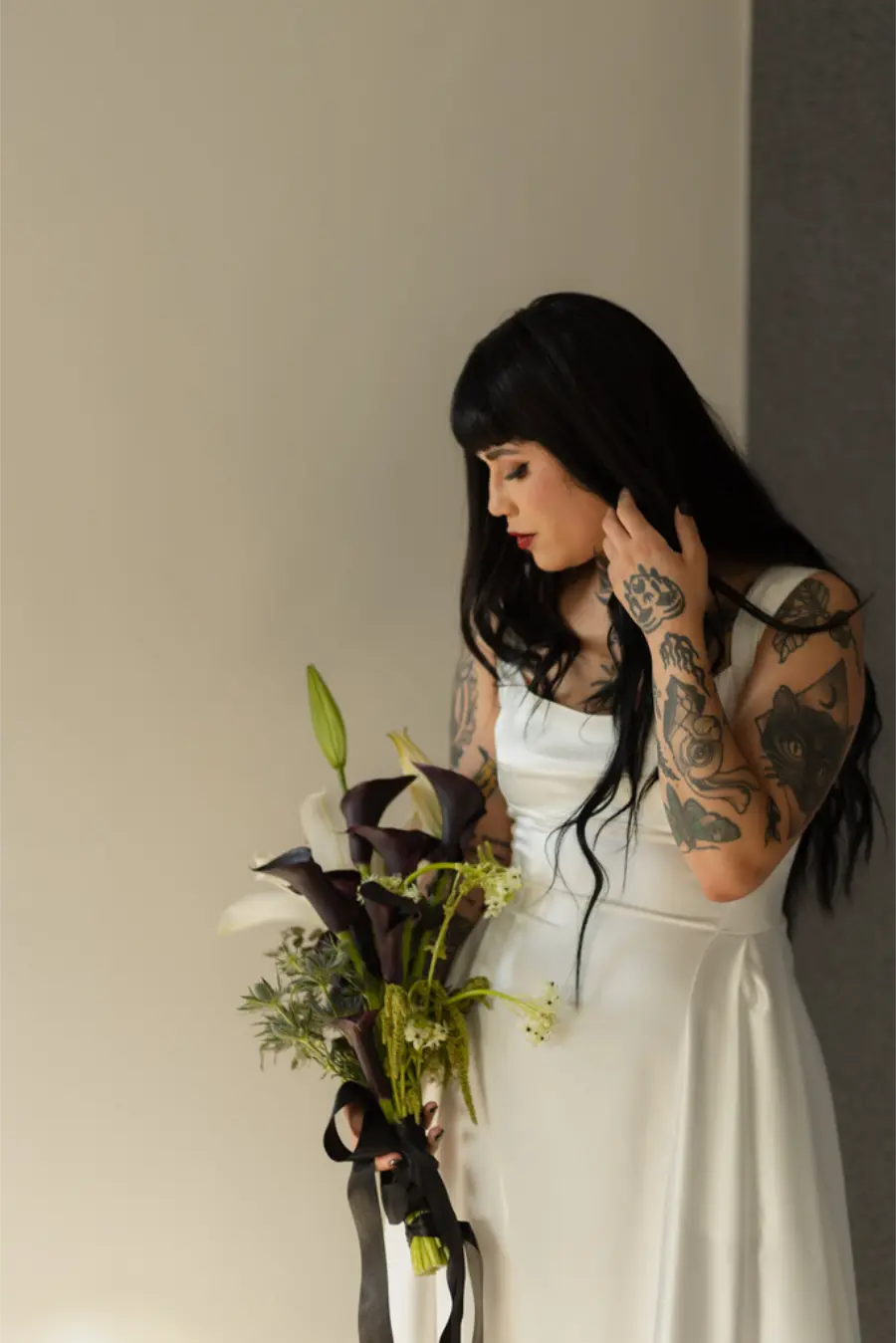 Noiva com tatuagens exibindo buqu&ecirc; minimalista de l&iacute;rios e flores escuras em contraste com seu vestido branco.