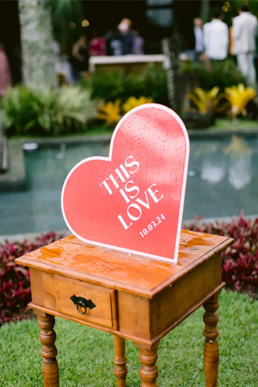 Placa em formato de coração vermelho com frase “This is Love” e data do casamento, apoiada sobre mesa de madeira em área externa.