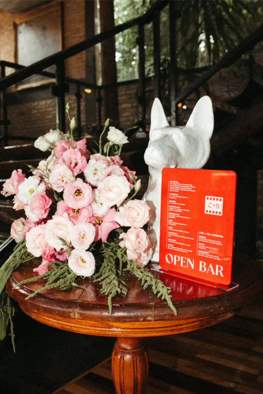 Arranjo de flores cor-de-rosa ao lado de placa vermelha de open bar personalizada para casamento.
