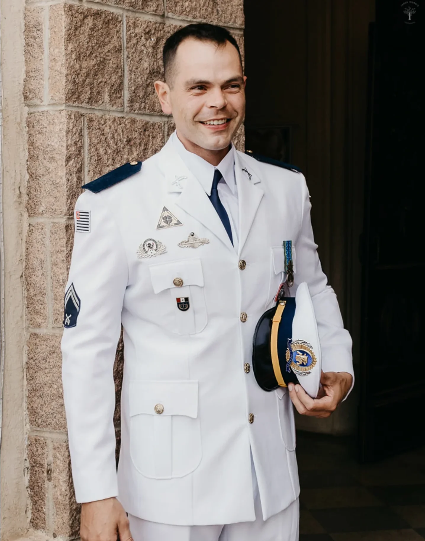 Militar em uniforme branco de gala sorri segurando o quepe durante cerim&ocirc;nia de casamento em frente &agrave; igreja.