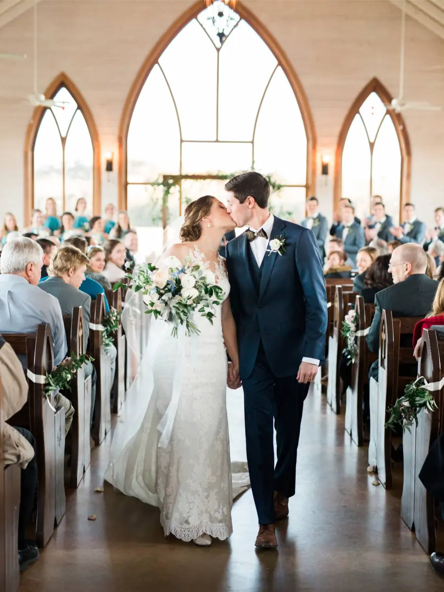Noivos se beijando ao caminhar pelo corredor de capela com vitrais e convidados aplaudindo