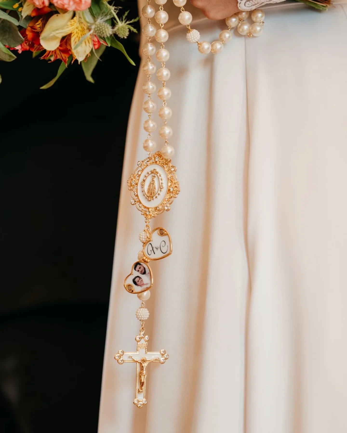 Terço de pérolas grandes com detalhes dourados e relicário com fotos pendurado junto ao buquê.