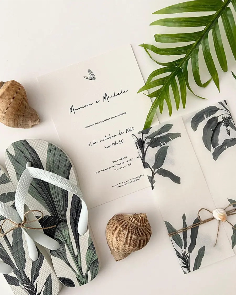 Convite de casamento tropical com folhas verdes, chinelo personalizado e envelope translúcido com estampa botânica, acompanhado de conchas e folhagem.