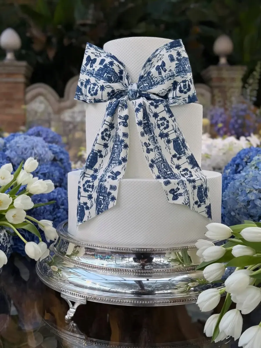 Bolo de casamento branco de três andares com laço estampado em azul inspirado em azulejo português, decorado com flores brancas e azuis.