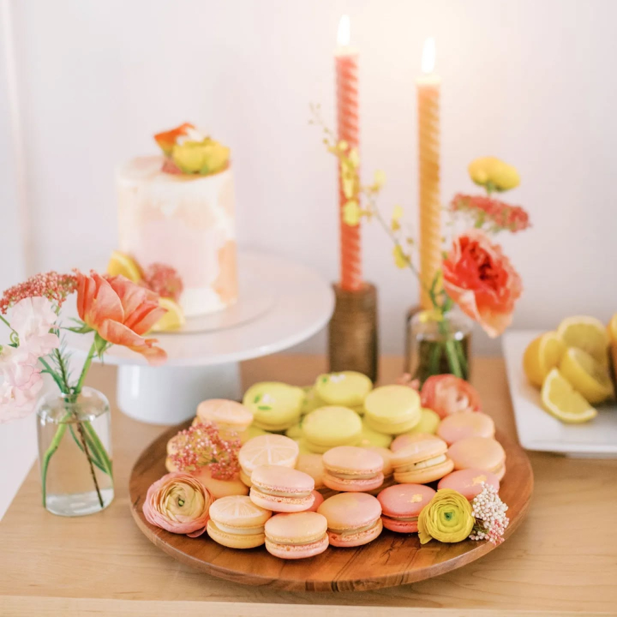 Prato com macarons amarelos e rosados, em meio a flores e decoração delicada, ao lado de um bolo minimalista e frutas cítricas.