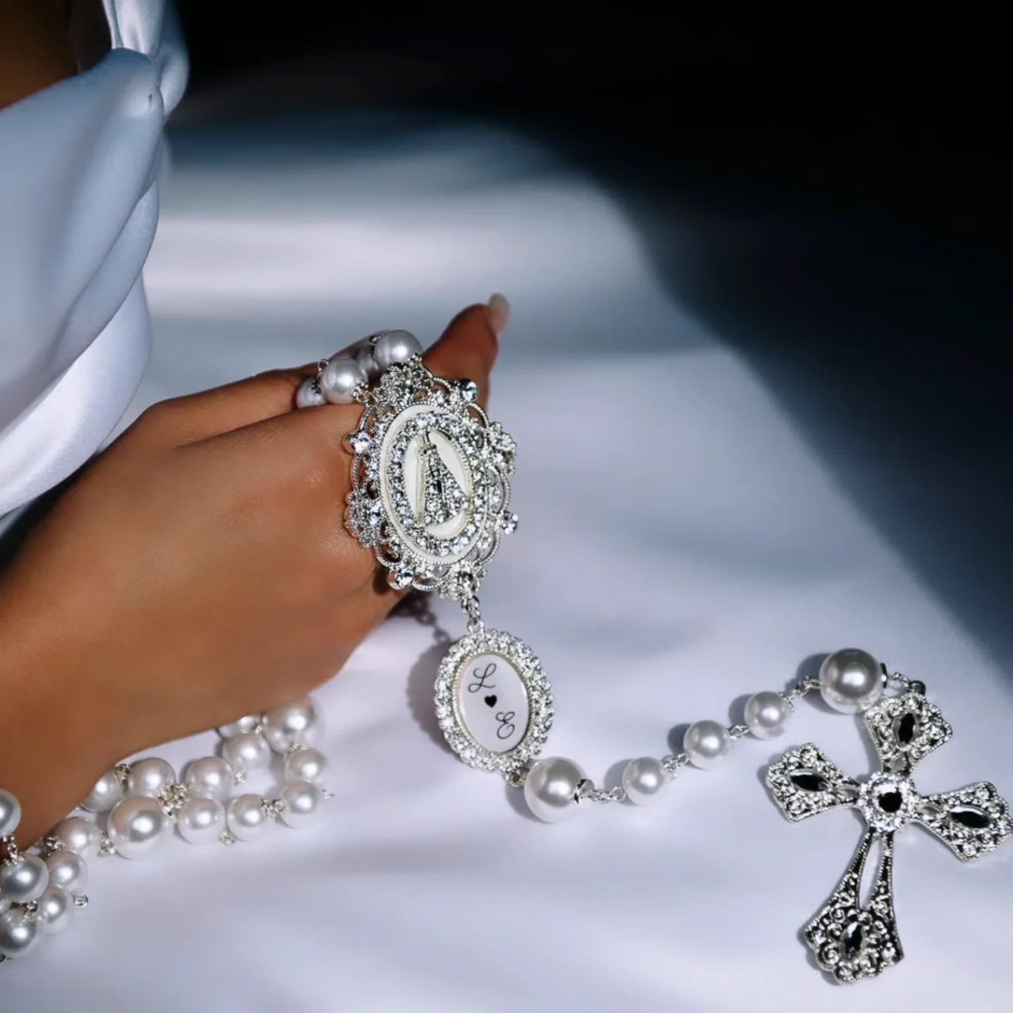 Noiva segurando terço de pérolas com relicário prateado em fundo branco.
