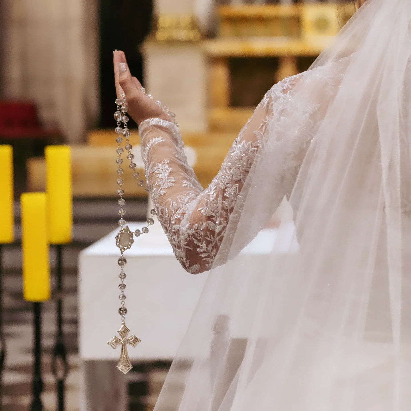 Noiva de costas segurando terço prateado em cerimônia religiosa.