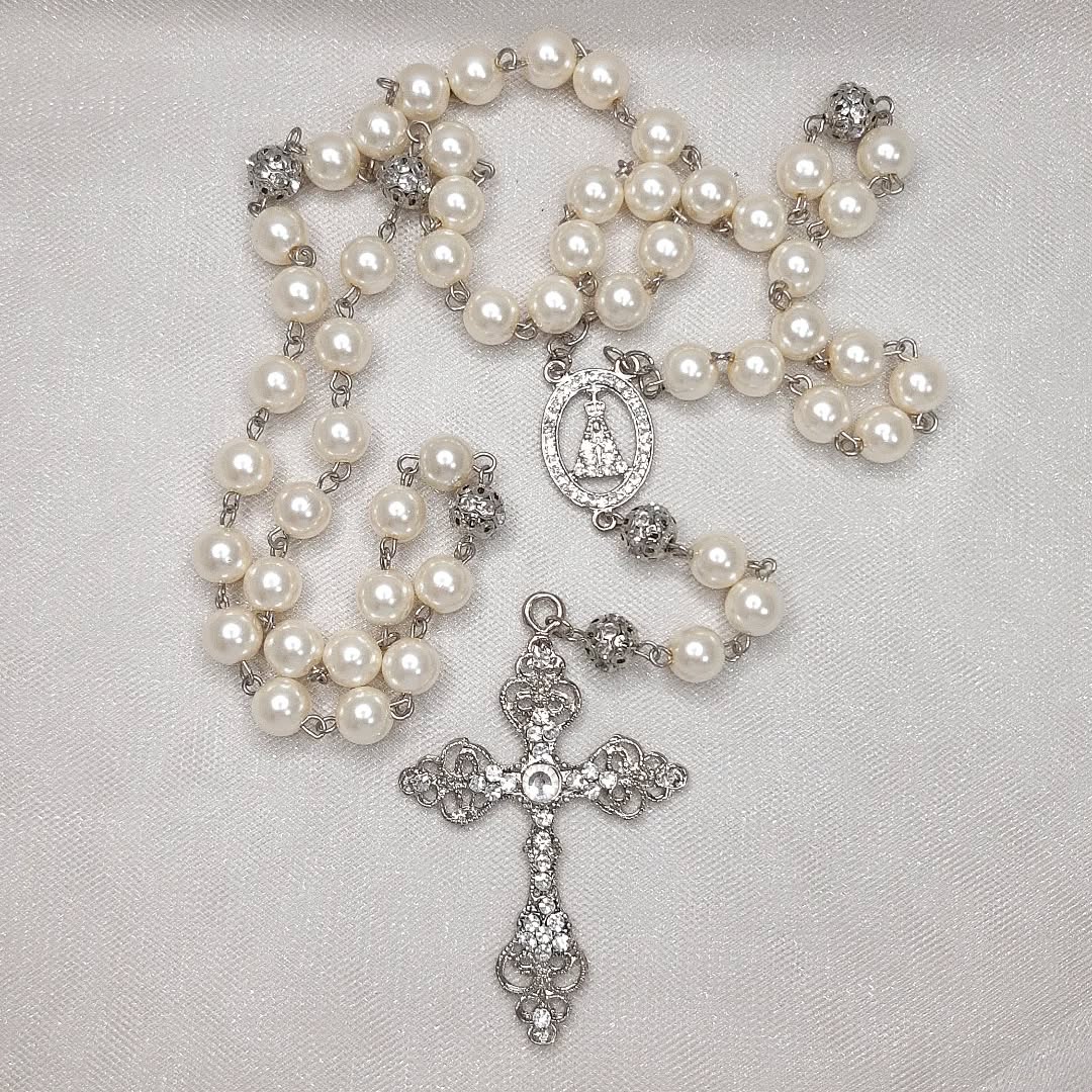 Terço prateado com contas peroladas e cruz decorada com detalhes em strass.
