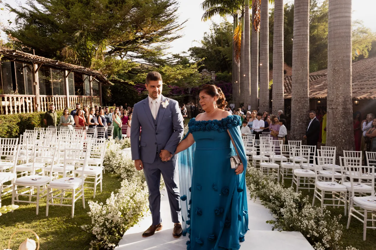 entrada do noivo de terno azul ao lado de m&atilde;e de vestido azul petr&oacute;leo