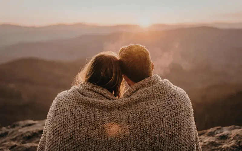 casal vendo o por do sol nas montanhas. Eles estão abraçados com uma manta de lã