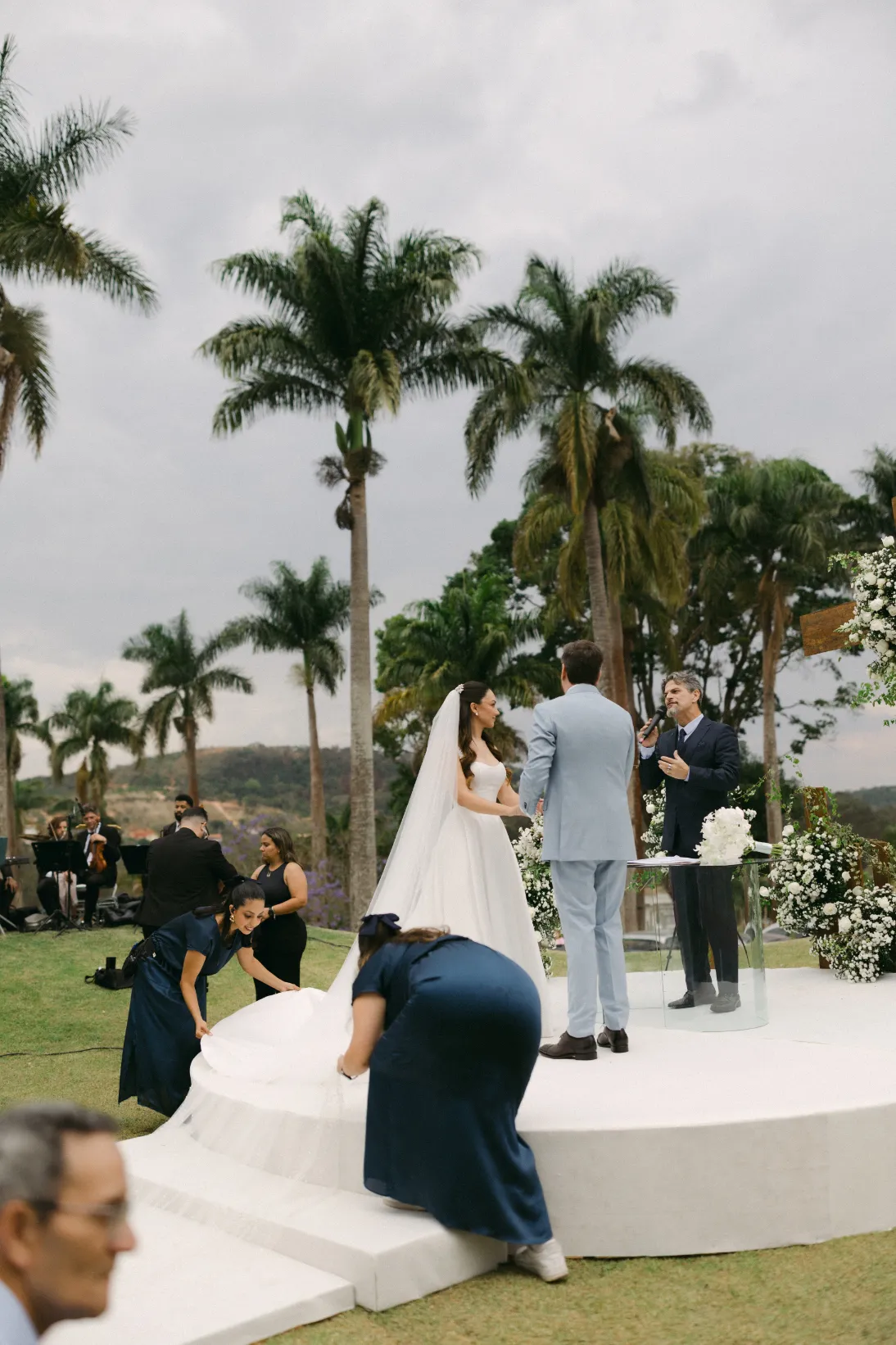 noivos no altar e assessoras segurando v&eacute;u