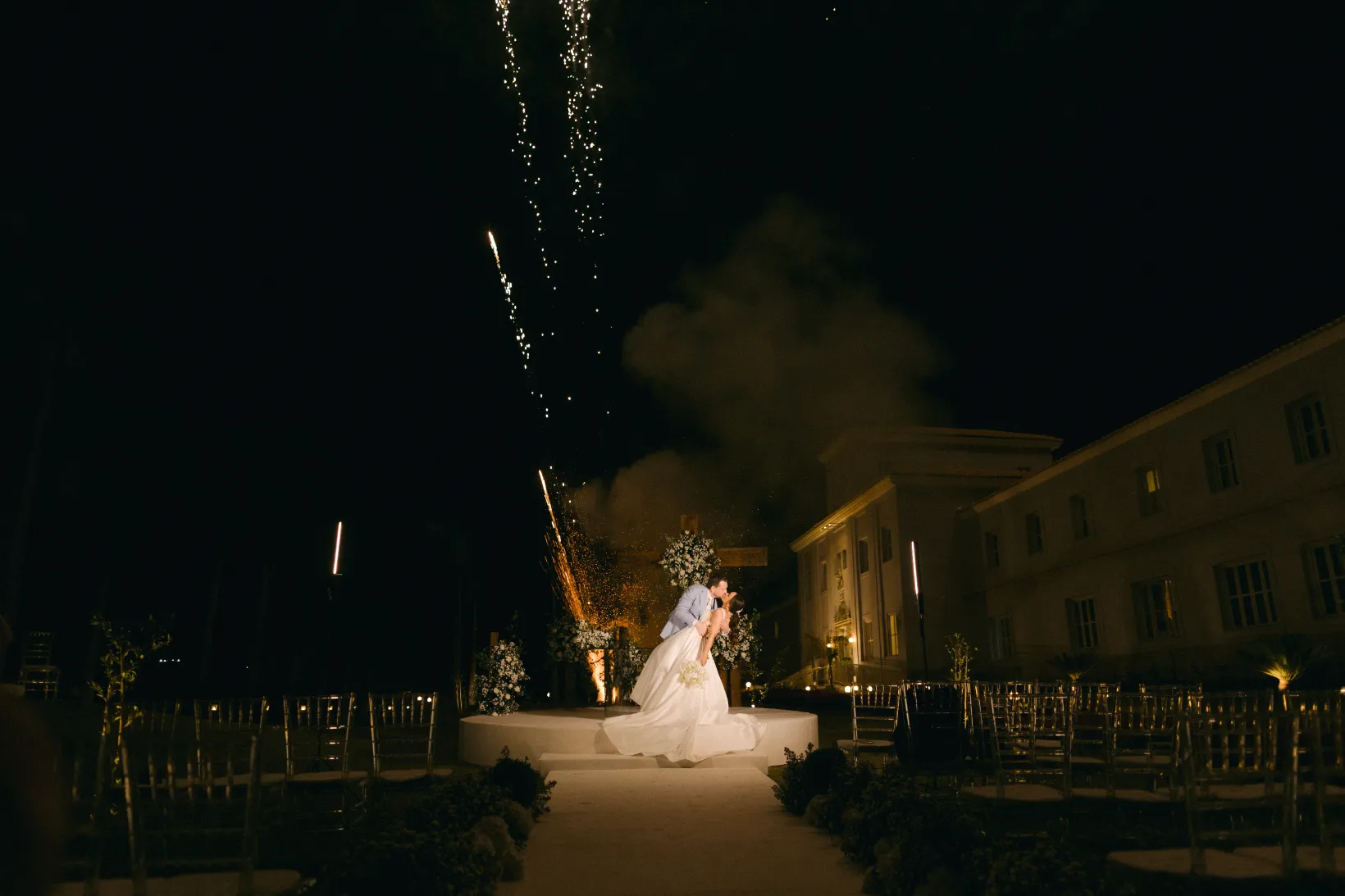 noivos se beijano enquanto s&atilde;o disparados fogos de artificio