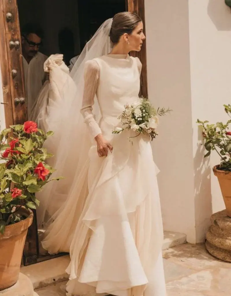 vestido de noiva estilo cl&aacute;ssico com mangas longas e saia em camadas. Ela est&aacute; saindo da igreja