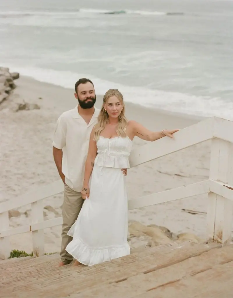 casal veste roupas brancas e beges claras, combinando com o ambiente praiano. A foto foi tirada em uma escadaria de madeira que leva à areia.