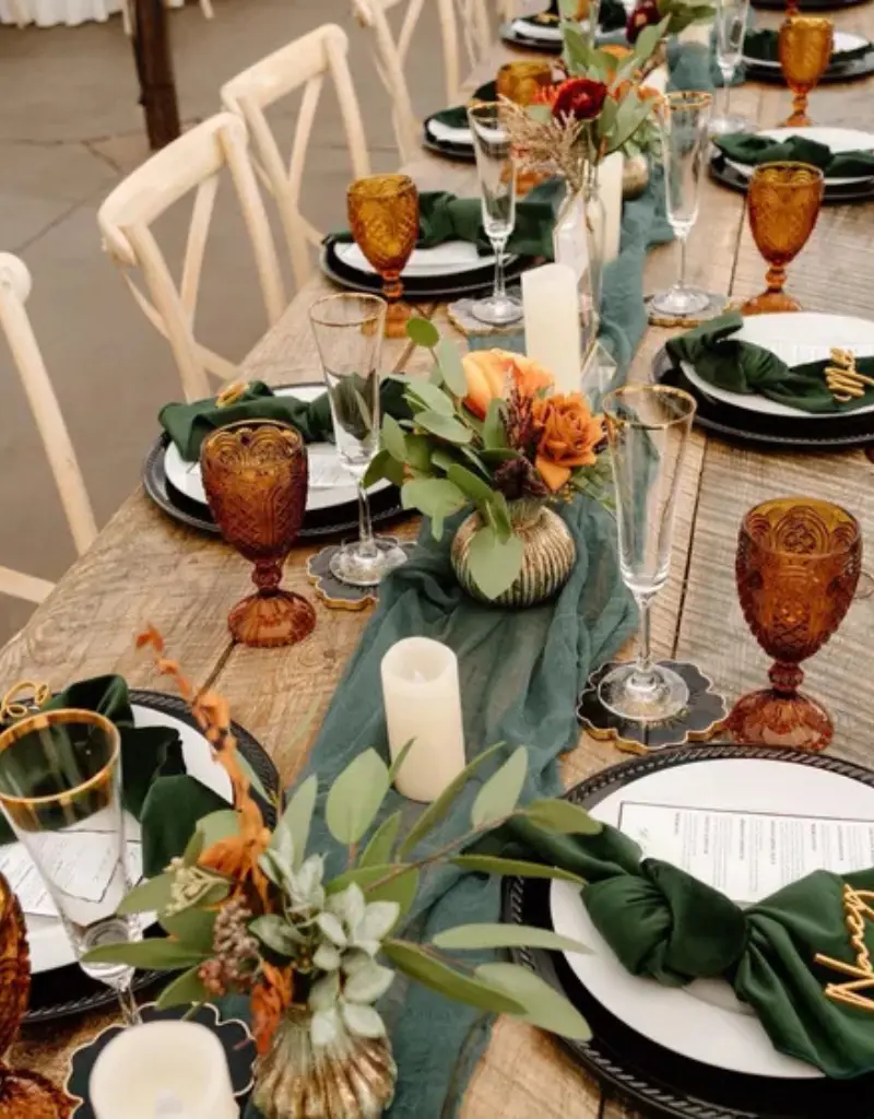 decoração de mesa posta com estilo rústico chic, apresentando um caminho de mesa verde sálvia, guardanapos verdes, taças âmbar e arranjos florais em tons de laranja